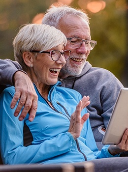 An older man and woman sitting outside on a bench looking at a tablet and smiling after seeing a Cigna insurance dentist in Virginia Beach
